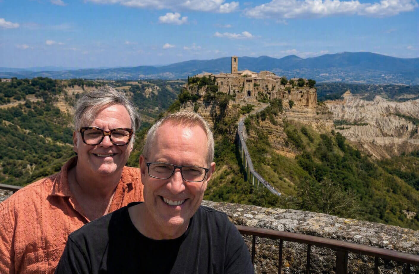 Two men smile for a photo in front of a hilltop village, overlooking a scenic landscape with mountains and blue skies in the background.