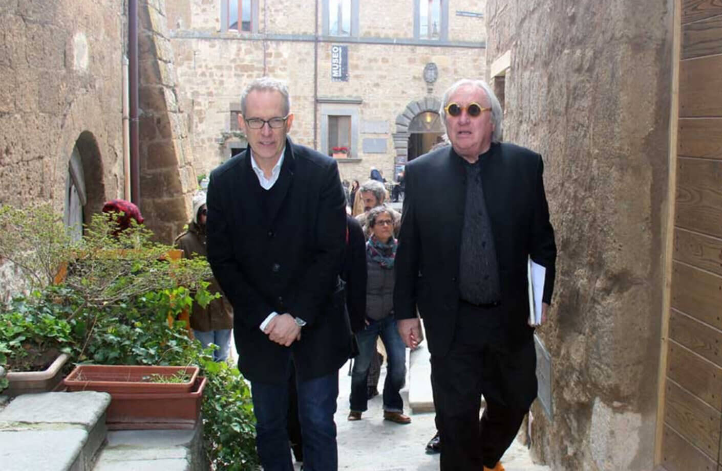 Two men walking down a narrow street framed by stone buildings, accompanied by a small crowd in the background.