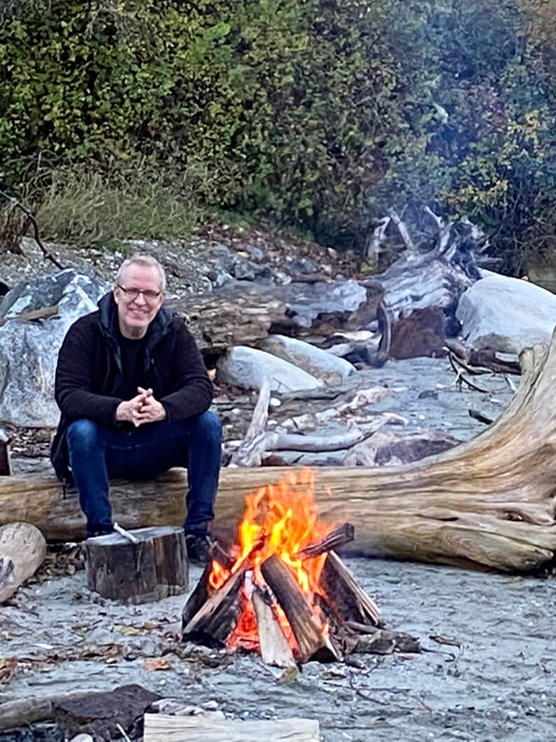 Man sitting by a campfire on the beach, surrounded by driftwood and rocks, with lush greenery in the background.