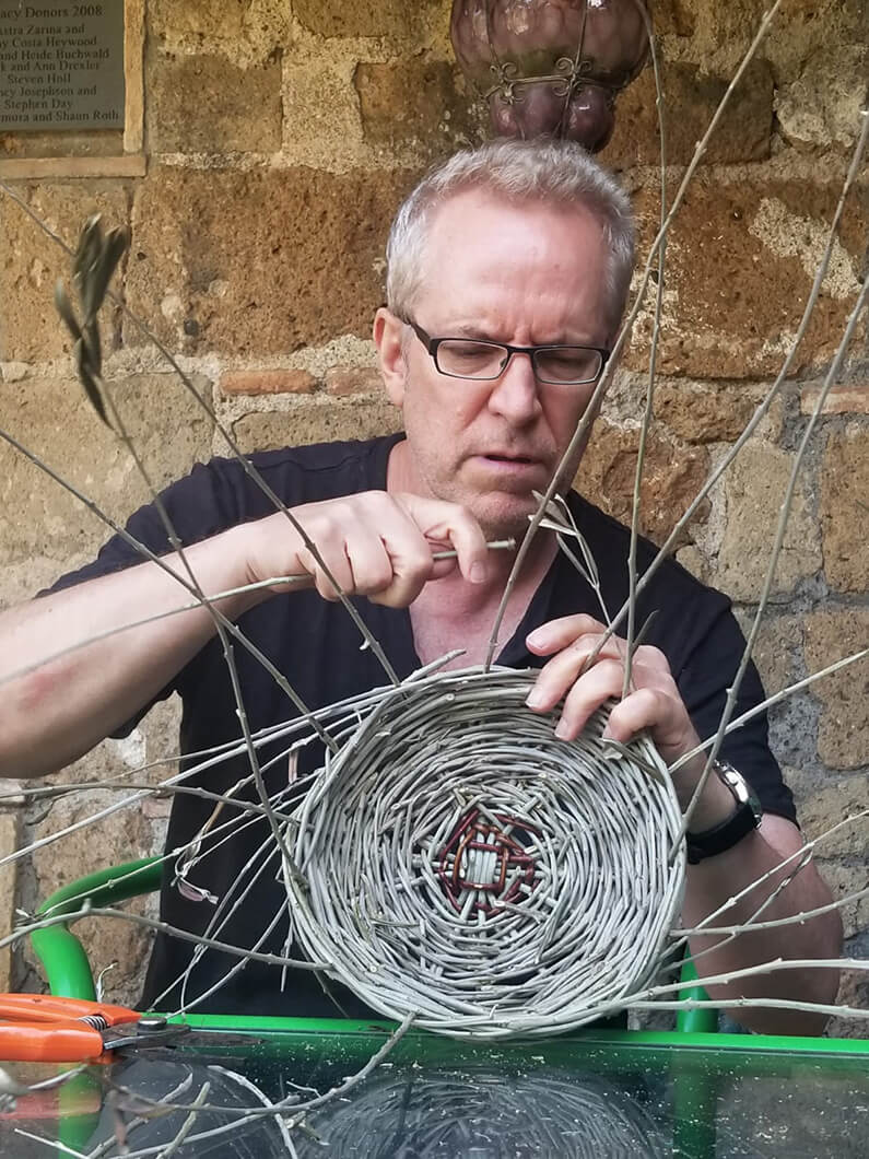 A craftsman weaves a basket with thin branches while focusing intently on his work. Tools are visible on a table nearby.