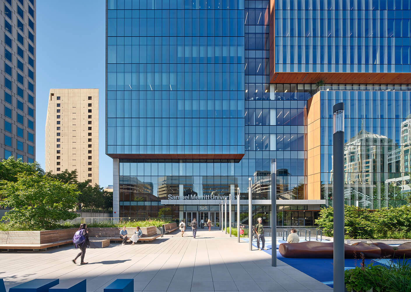 Front view of the Samuel Merritt University building, showcasing glass panels and landscaped seating area. People are walking and relaxing outside.