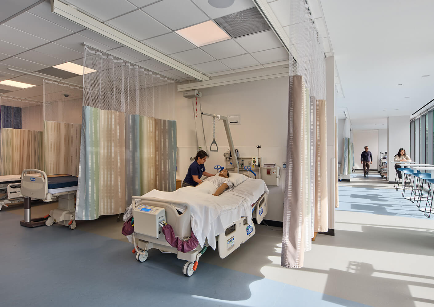 A hospital room with a patient in bed receiving care from a nurse, surrounded by privacy curtains and medical equipment.