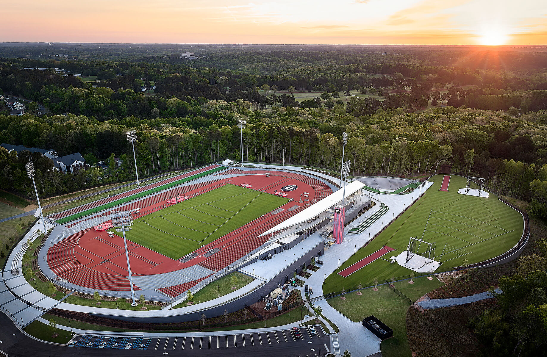 Aerial view of a sports complex featuring a track and field area, grass fields, and surrounding trees during sunset. Parking and facilities visible.