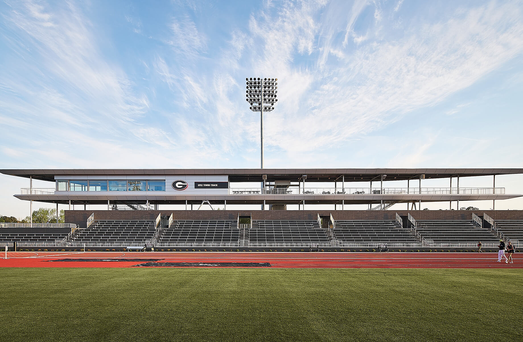 Stadium view with bleachers and a press box, featuring a track and field area under a clear sky with athletic lighting.