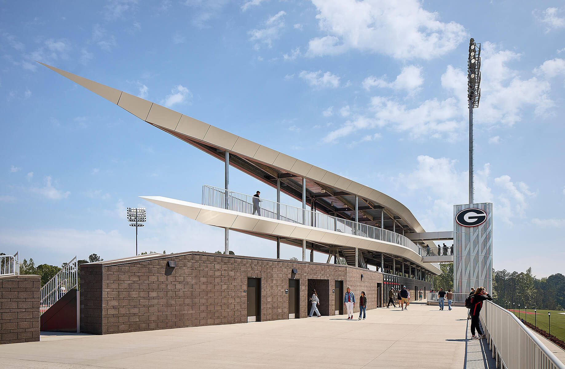 Stadium with a unique roof design, featuring a viewing deck and accessible entrances. Clear blue sky and people present on-site.