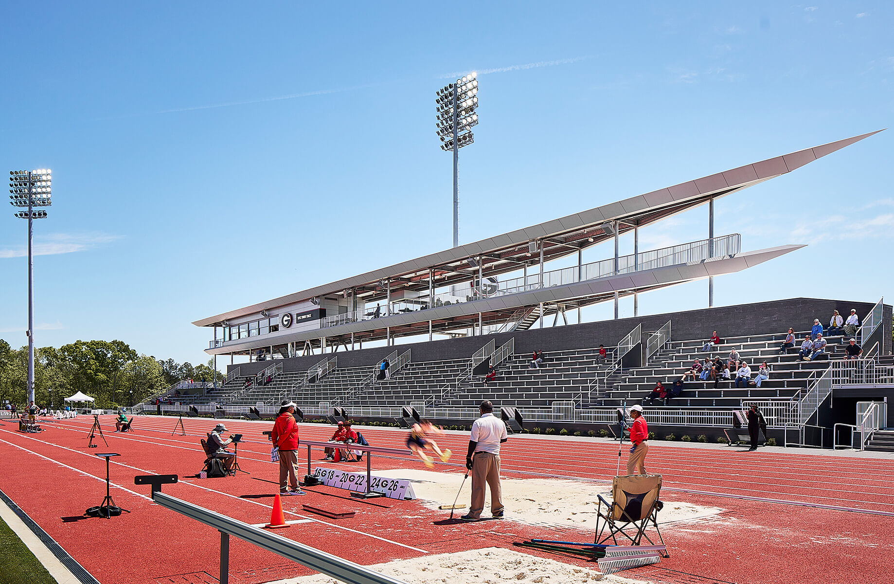 Athletic field with red track and long jump area, featuring a large spectator grandstand and floodlights under a clear blue sky.