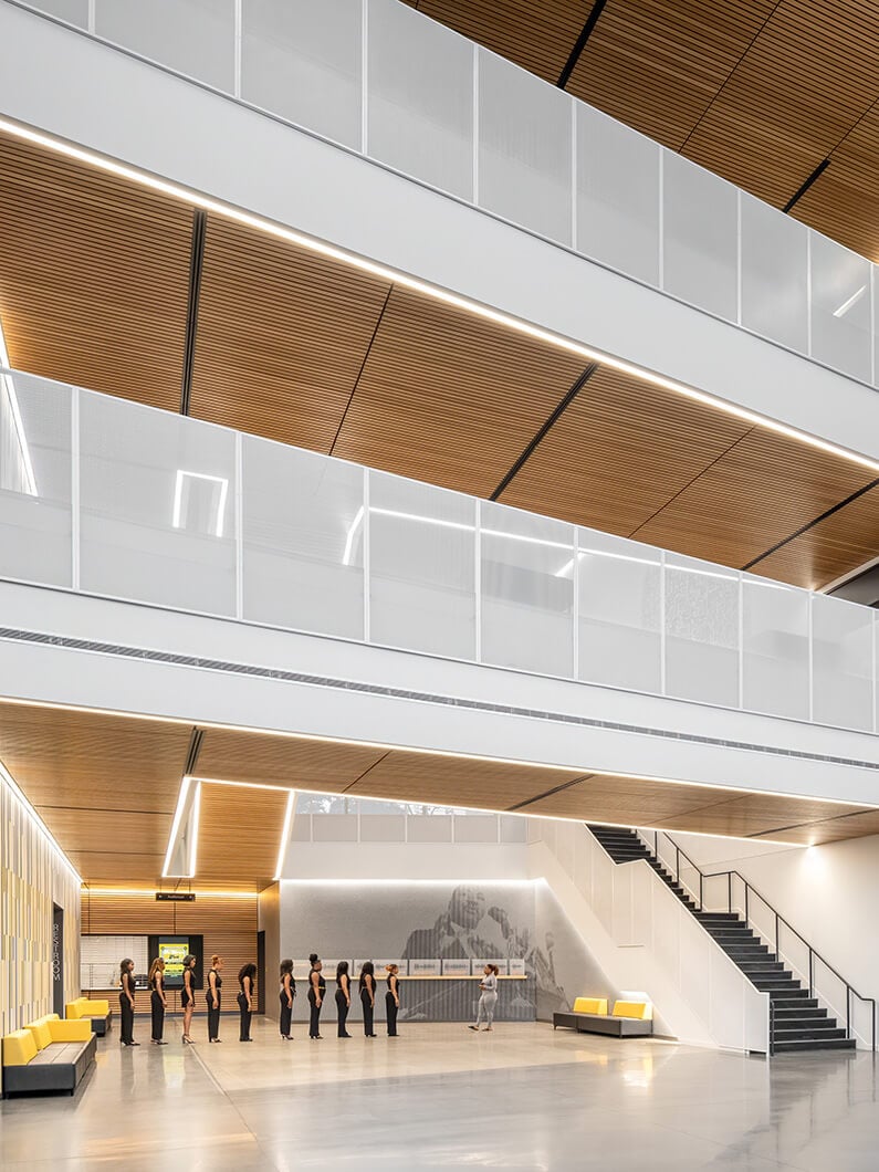 Interior view of a spacious atrium with wooden ceilings and sleek lighting. A line of people dressed in black stands on polished concrete flooring.