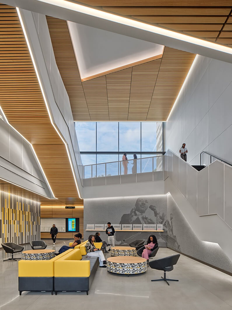 Interior space with seating area, large windows, and wooden ceiling. People engage in conversation and leisure activities. Staircase in the background.