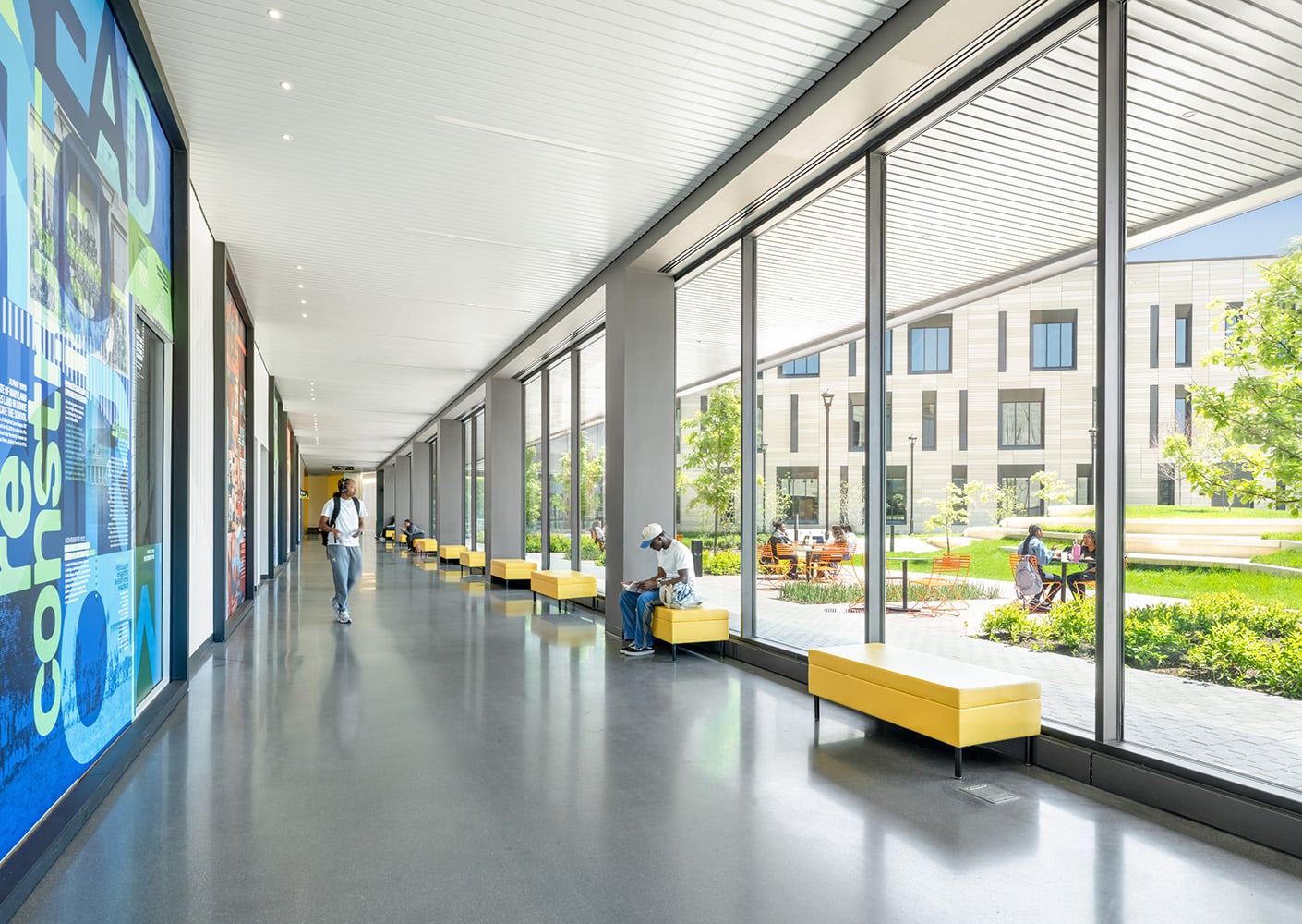 Interior hallway with large windows, yellow benches, and artwork, showing a person walking. Lush greenery and gathering area visible outside.