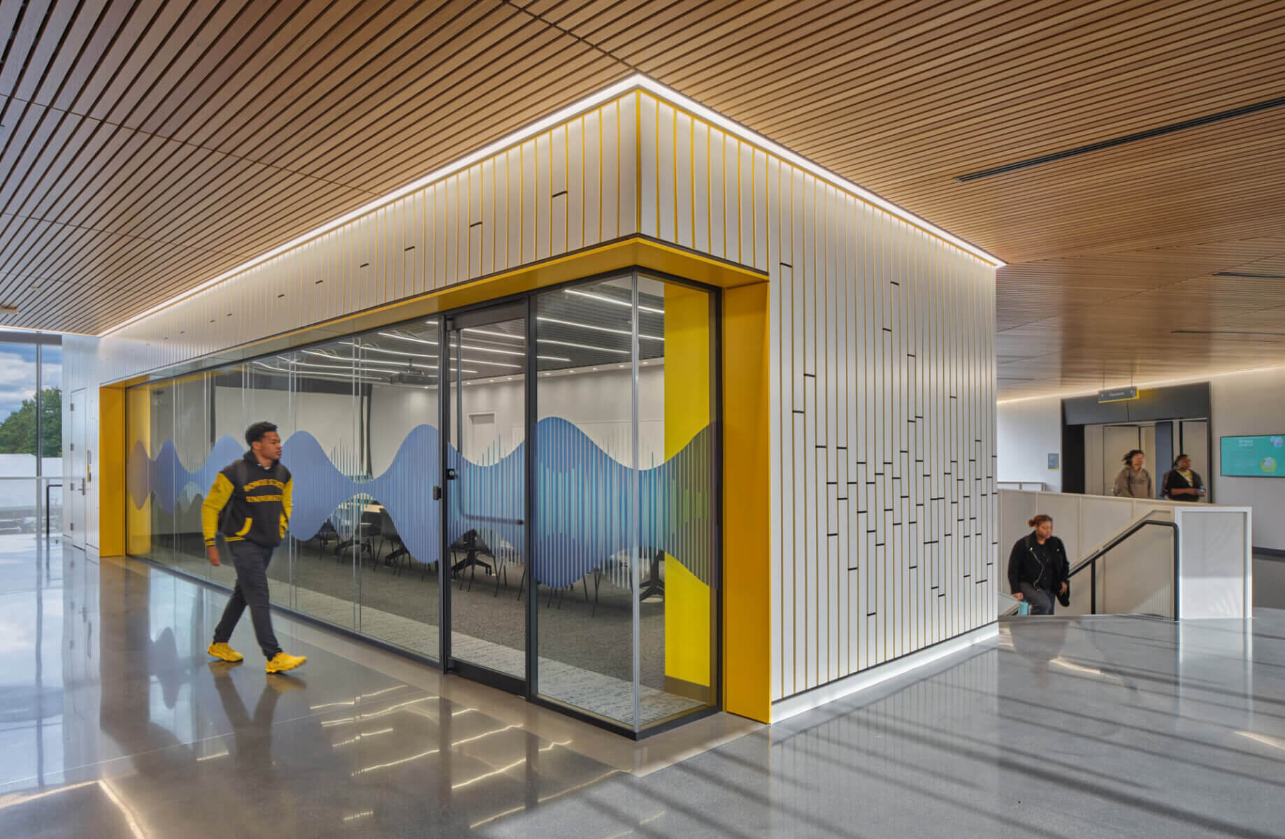 Interior view of a campus building featuring a glass-walled conference room, sleek lines, yellow accents, and people walking by.
