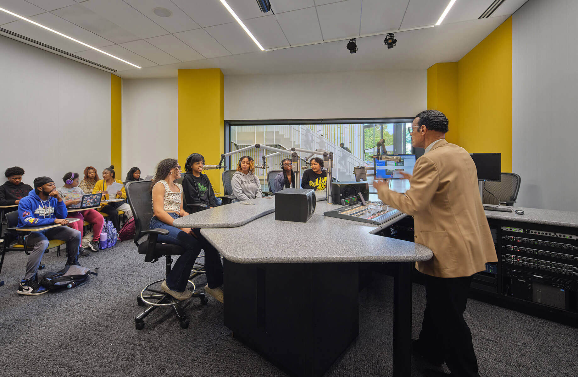 Broadcast studio scene with an instructor speaking to students seated at desks. Digital equipment and soundproofing visible in background.