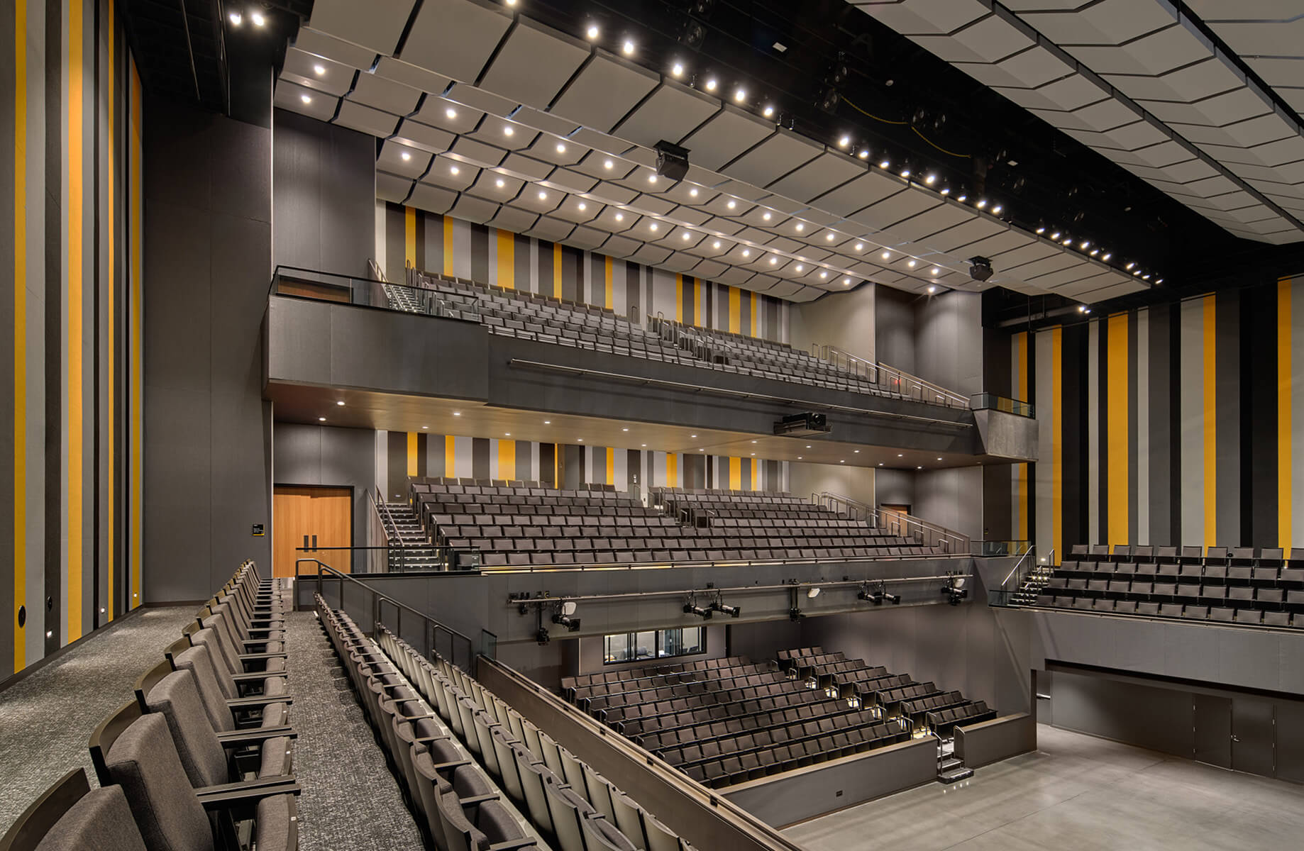 Interior view of a theater with tiered seating, featuring yellow and gray striped walls and stage lighting. Empty seats arranged for performances.