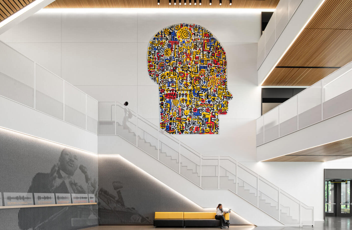 Colorful mural of a human head silhouette on a white wall in a spacious interior, with a person sitting on a yellow and black bench.