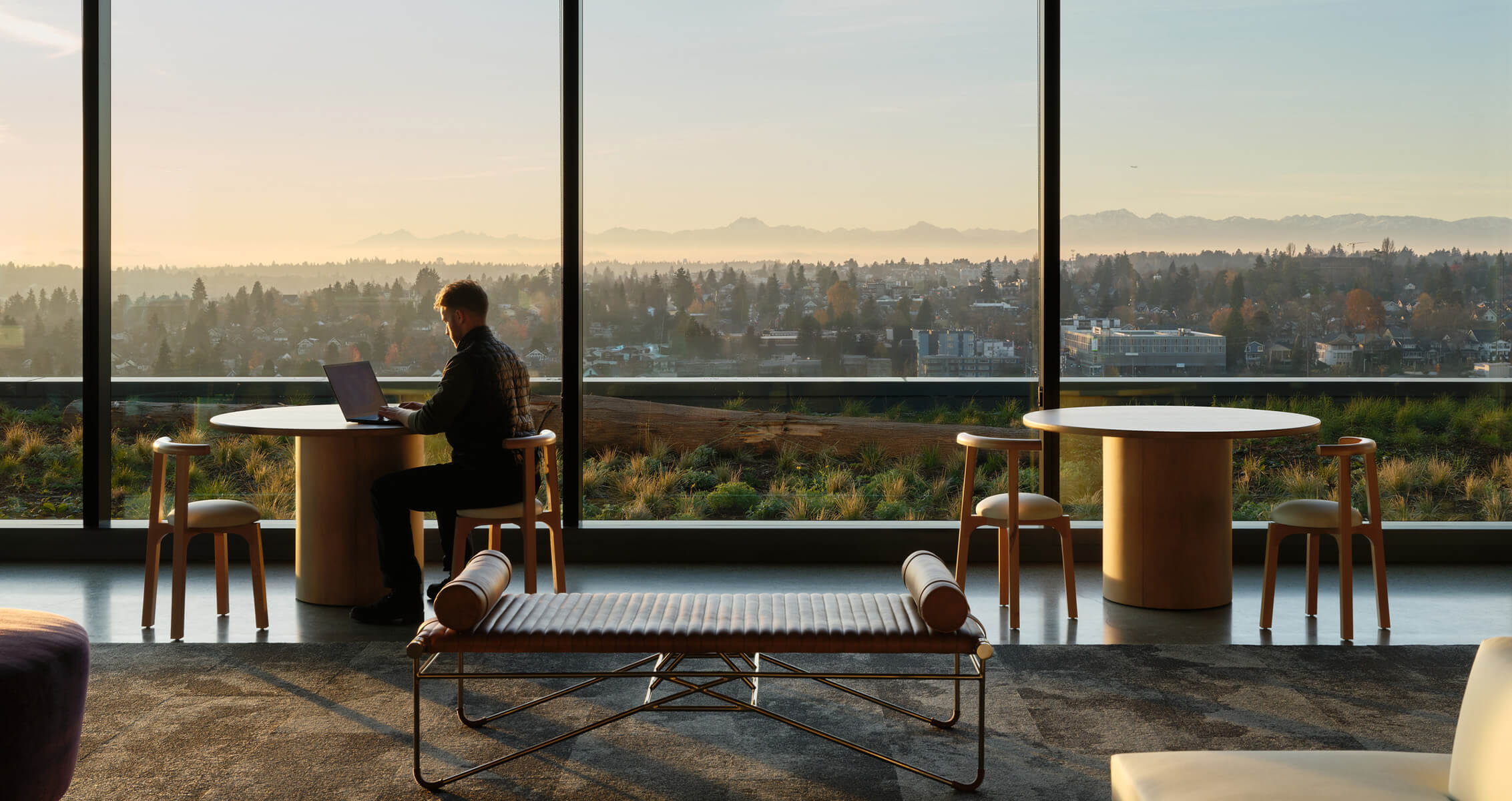 Person working on a laptop at a table, with a view of a city skyline and mountains through large windows. Comfortable seating in the foreground.