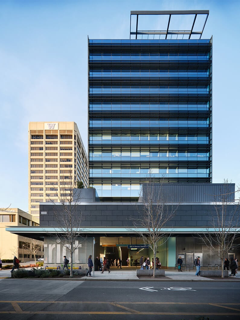 Glass-fronted building with multiple stories, featuring a large entrance area and pedestrians in front, with a taller building in the background.