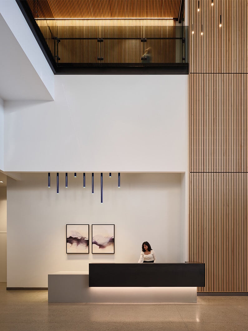 Reception area featuring a sleek front desk, a woman attending, and framed artwork on a wall, paired with wood paneling and modern light fixtures.