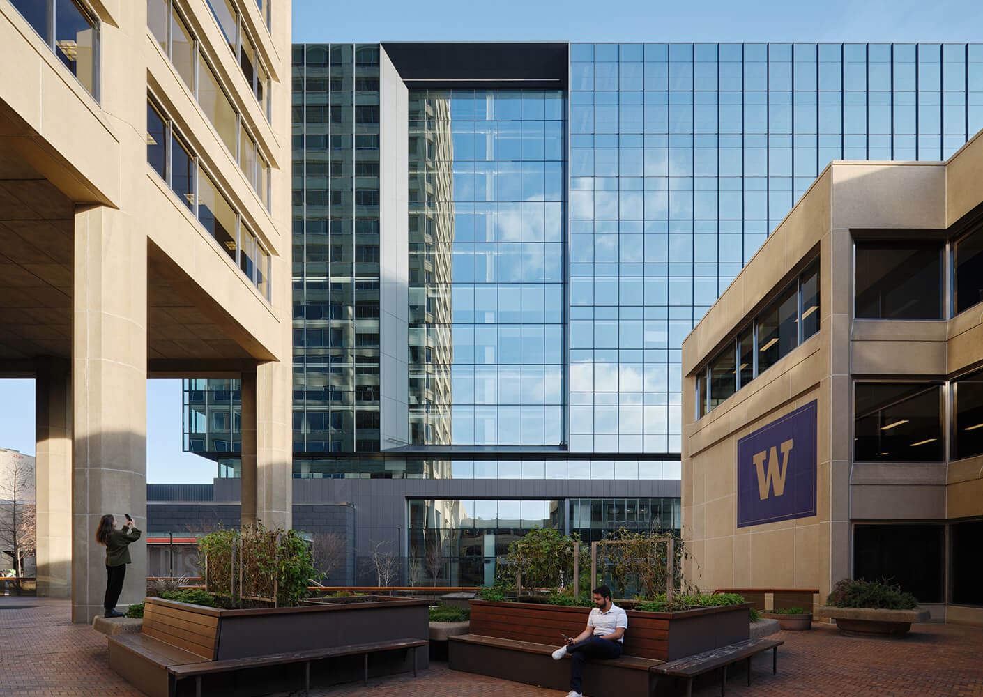 A courtyard scene featuring two buildings with large glass facades. A person sits on a bench while another takes photos nearby, surrounded by plants.