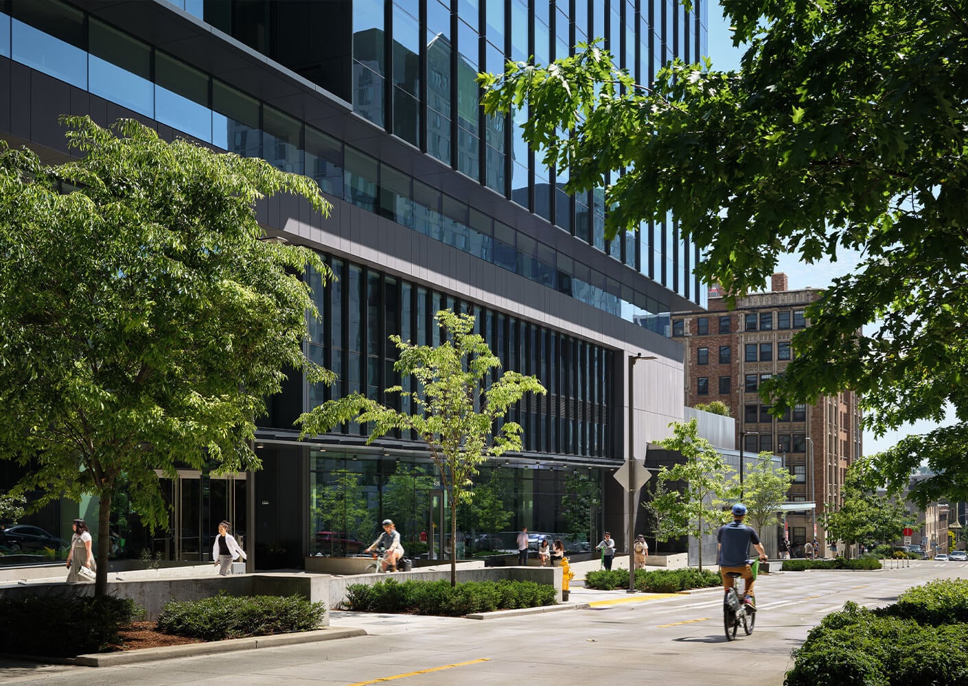 Urban streetscape featuring a glass building, green trees, and pedestrians, with a cyclist in a city setting. Clear skies above.