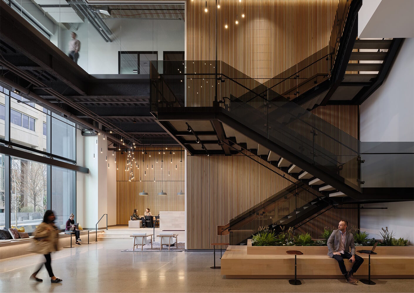 Alt text: Interior view of a spacious lobby featuring a staircase, seating areas, wooden paneling, and large windows with natural light.