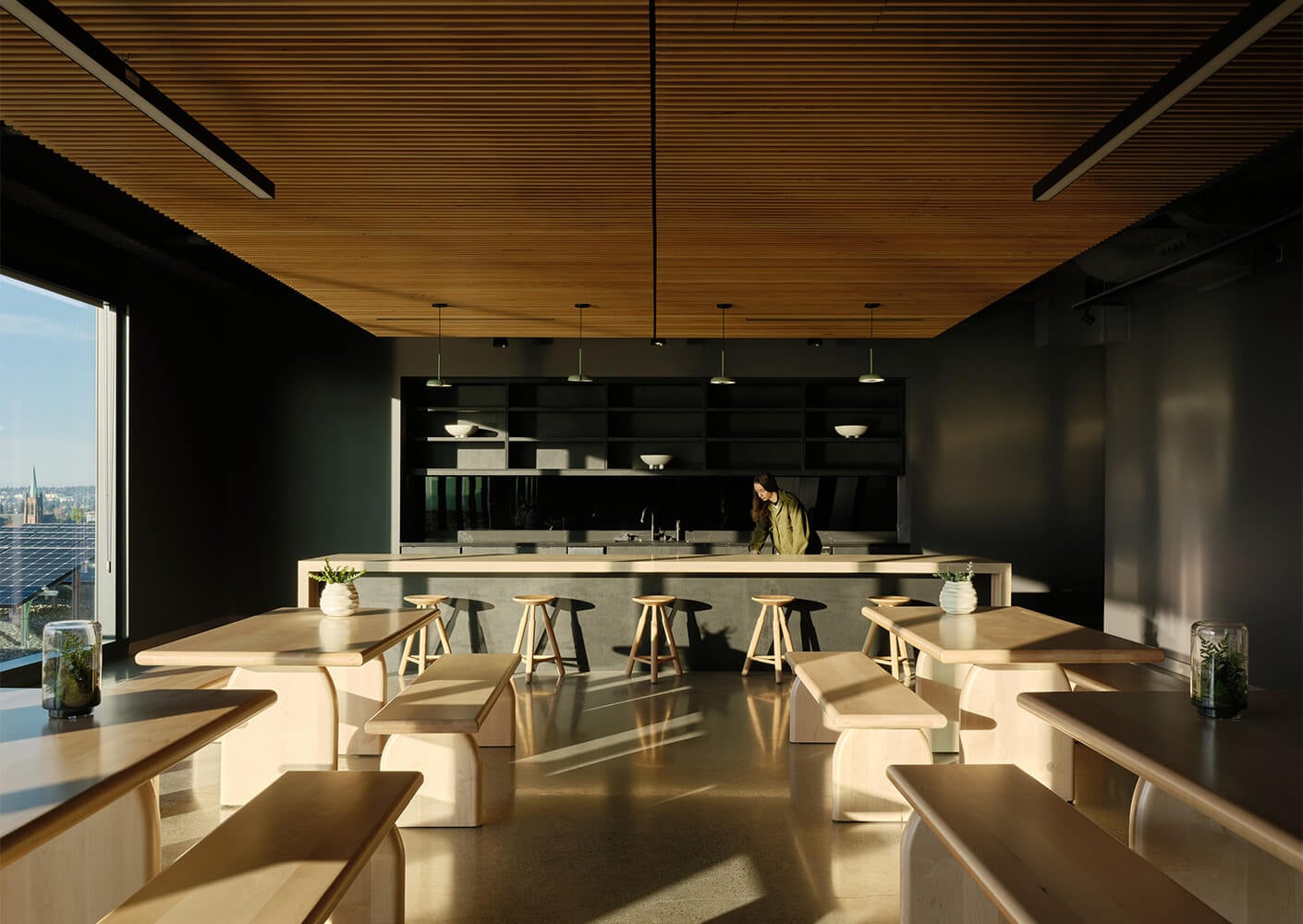 A spacious interior with wood elements, featuring tables and stools. A woman stands at a counter in front of large windows.