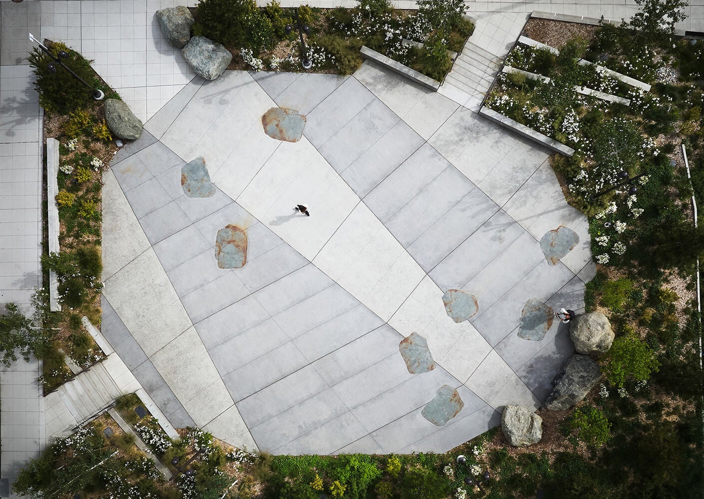 Aerial view of a concrete plaza with irregular stone placements and surrounding greenery, featuring a person walking through the area.