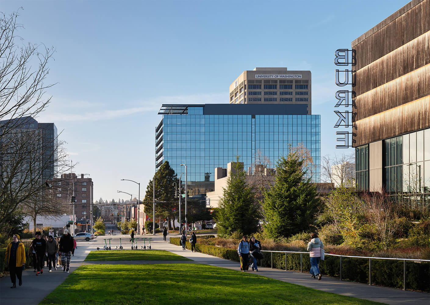 Urban scene featuring pedestrians on a sidewalk, green spaces, and various buildings, including the University of Washington and Burke Museum.