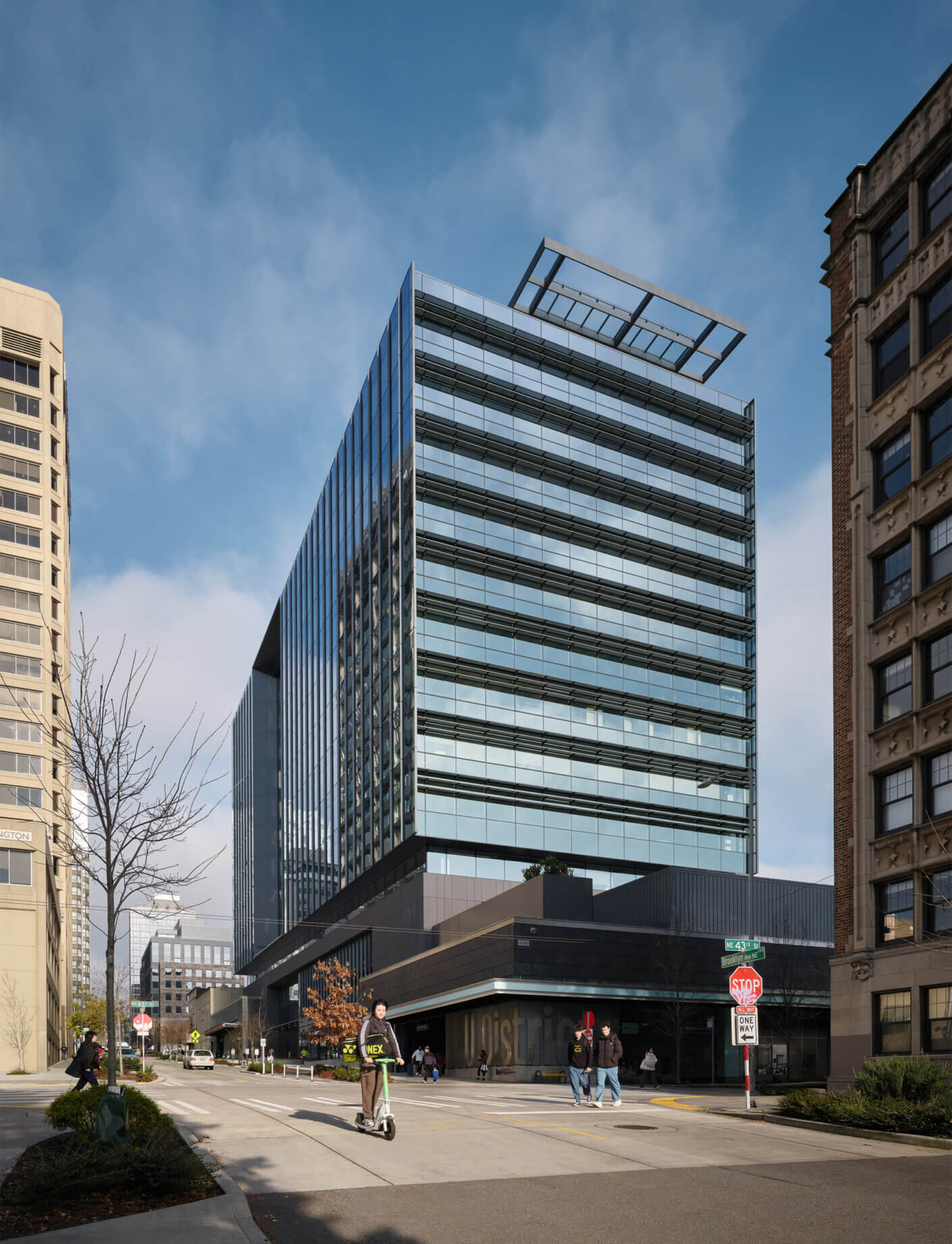 City building with glass facade and distinct architectural features, surrounded by pedestrians and urban streets. Blue sky overhead.