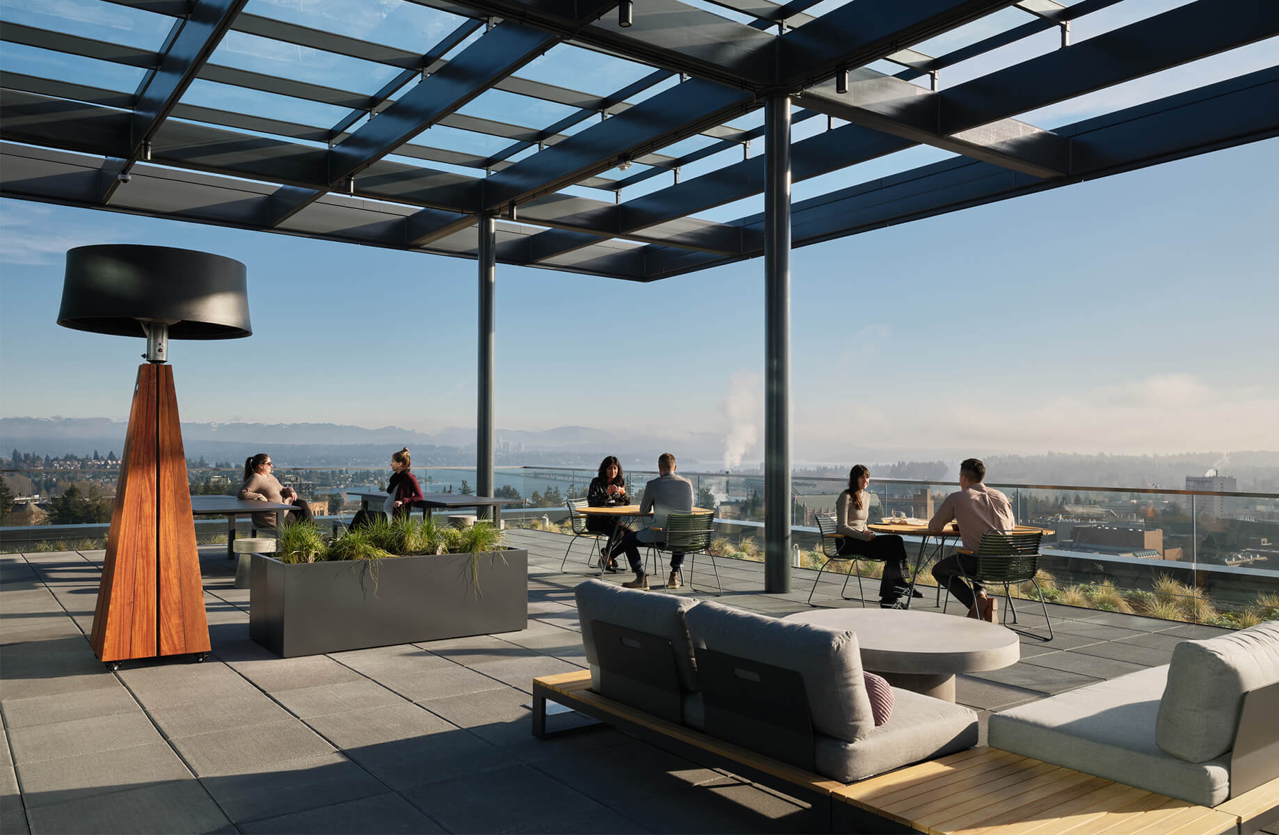 Rooftop space with seating areas, planters, and a lamp. People work and socialize against a backdrop of mountains and city views.