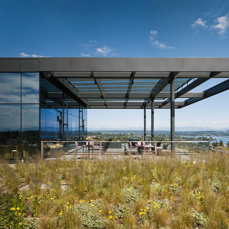 Rooftop terrace featuring glass walls and metal framing, with seating and greenery against a backdrop of mountains and sky.