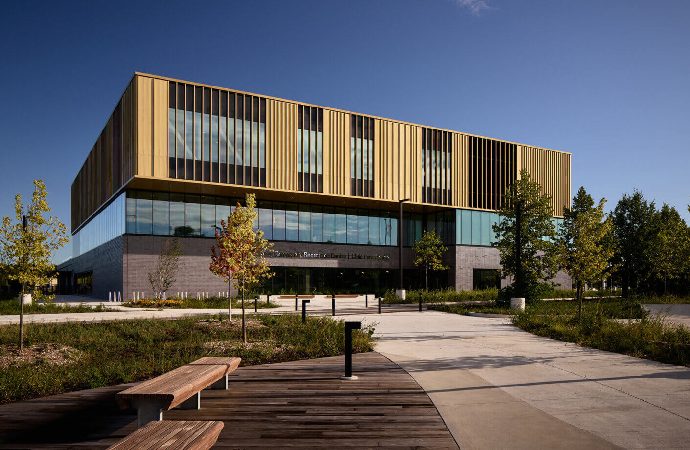 Exterior view of a sleek, multi-level community center featuring large windows, a contrasting gold façade, and landscaped grounds with pathways and seating.