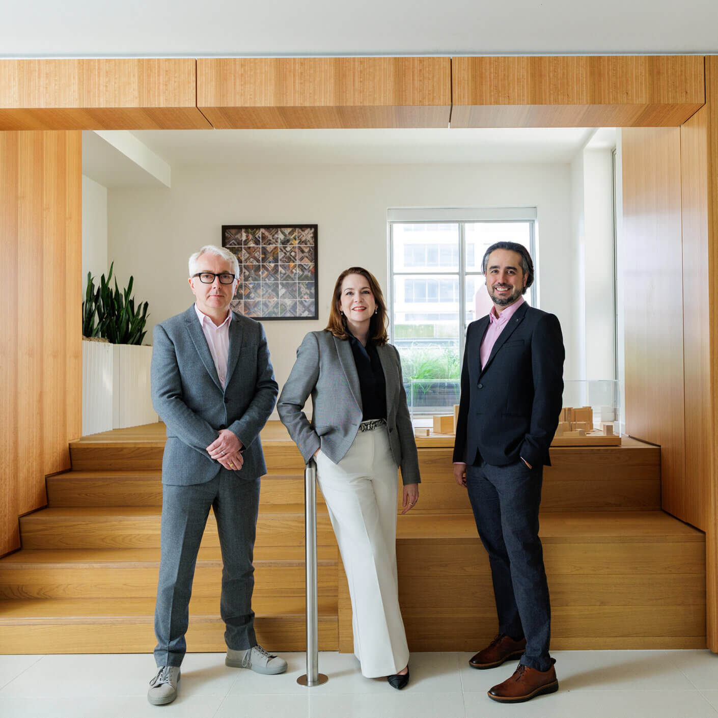 Three professionals dressed in suits stand together in an office space, with wooden accents and architectural models in the background.