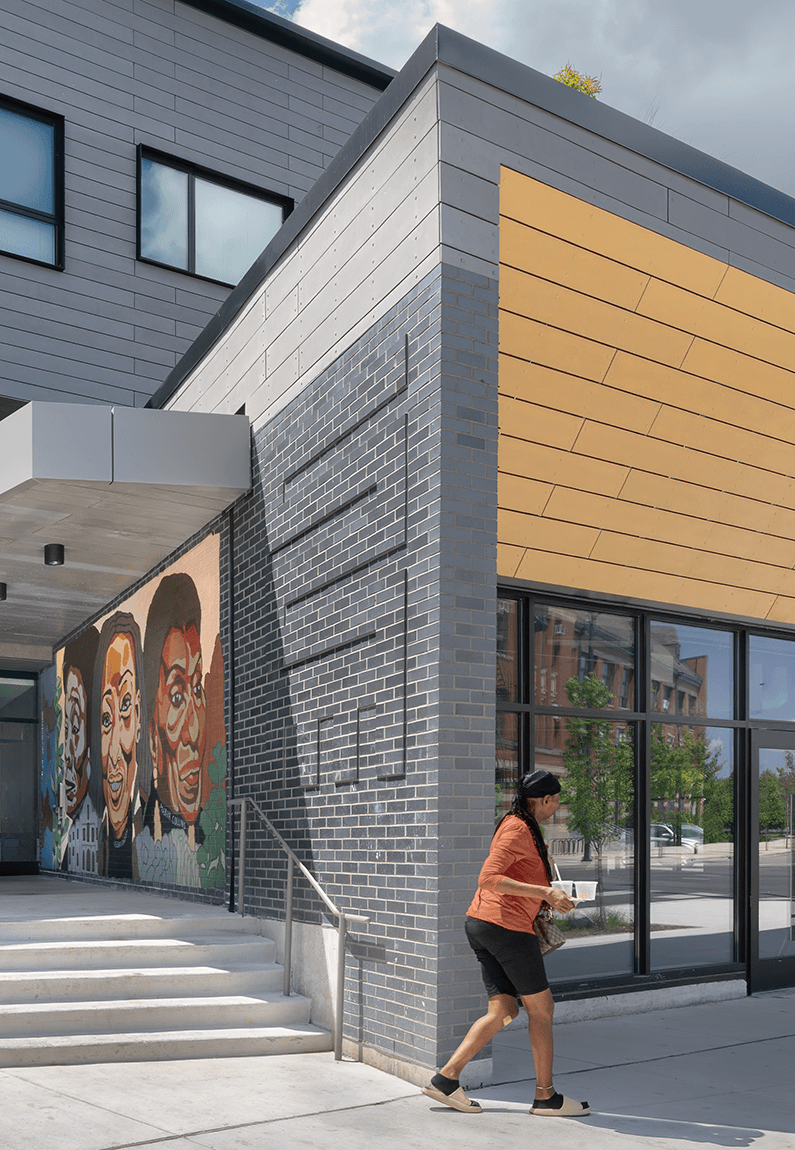A person walks by a building with a gray and yellow facade, featuring a colorful mural of faces on one side. Steps lead to the entrance.