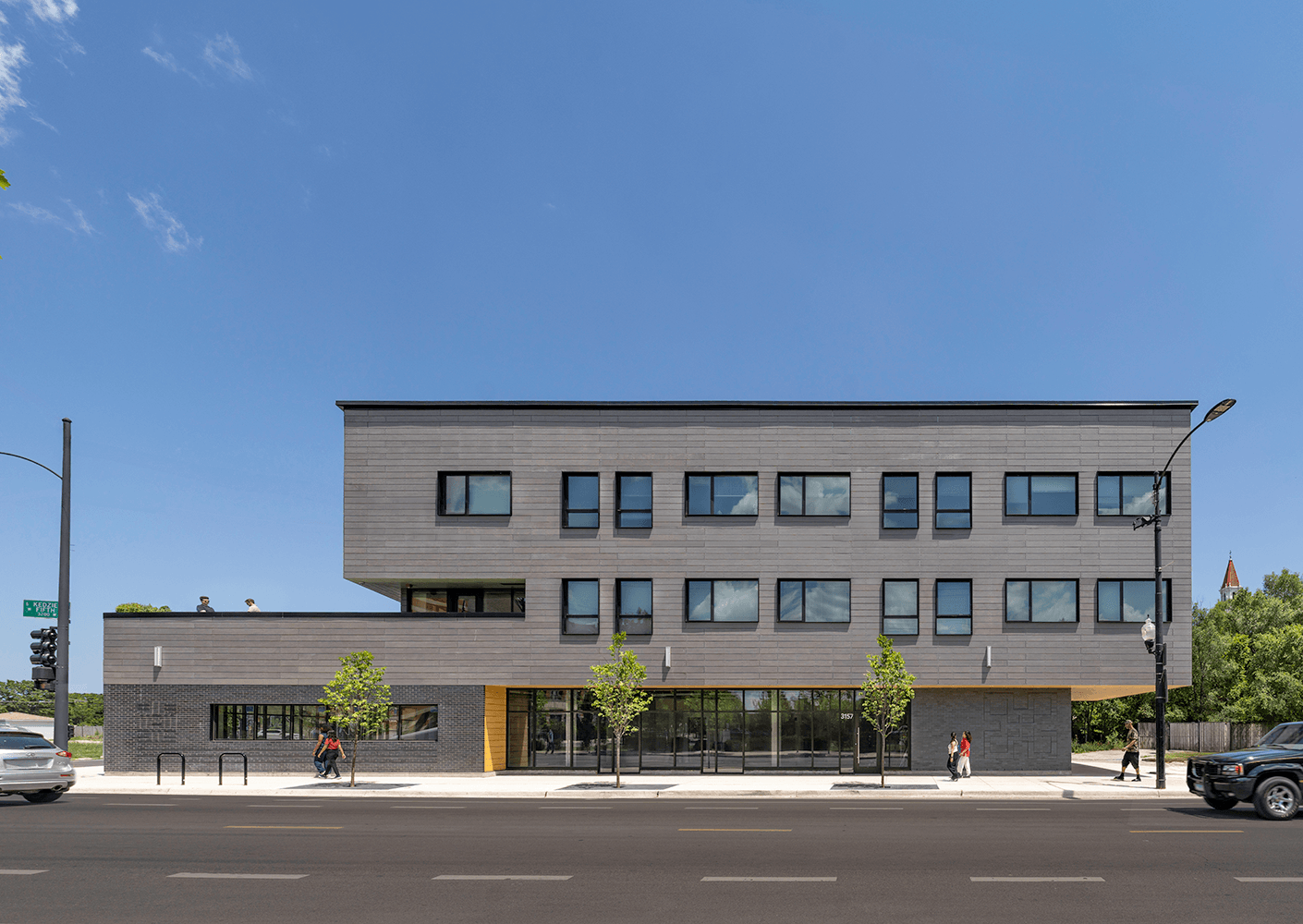 Contemporary three-story building with large windows, flat roof, and green landscaping along the street. Pedestrians walk past on sidewalk.