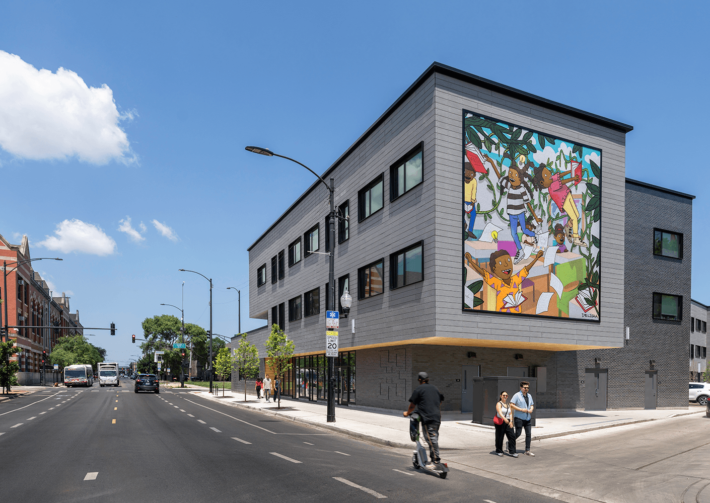 Urban building with mural depicting children playing, large windows, and street activity. Clear blue sky overhead and traffic visible.