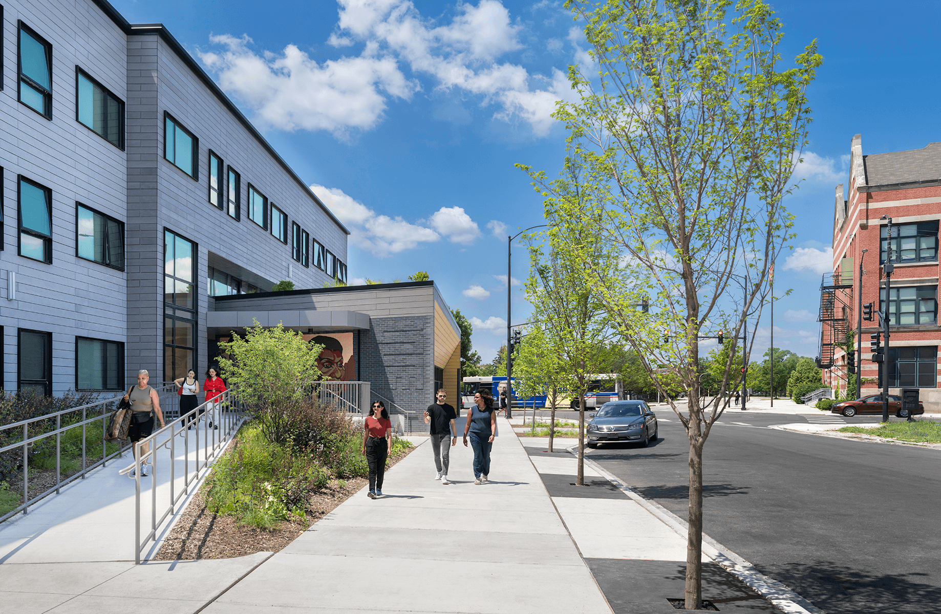 People walking along a sidewalk near a contemporary building with large windows and landscaping. Clear blue sky enhances the scene.