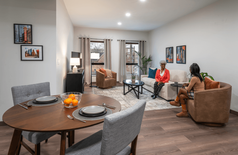 Two women converse in a spacious living area featuring a dining table, seating, and artwork on the walls. Natural light brightens the room.