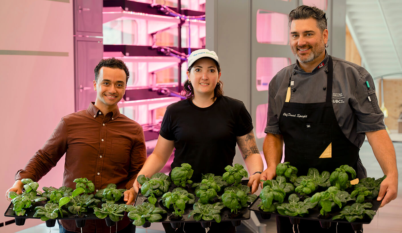 Three individuals hold trays of fresh green plants in a bright indoor setting, with vertical farming equipment in the background.