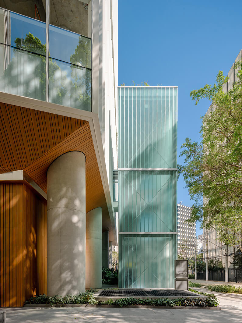 Glass and concrete building structure with wooden accents and landscaped areas, featuring large windows and a clear blue sky.