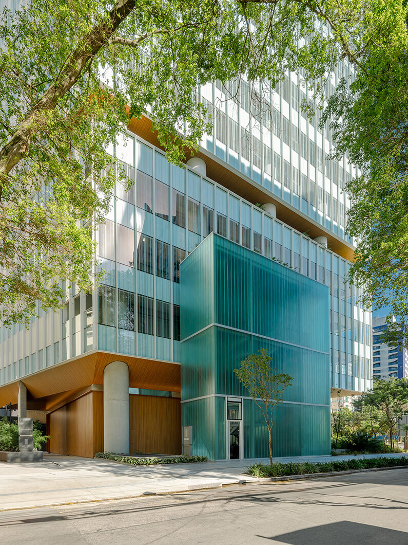 Glass and wood building exterior with unique teal section surrounded by trees and landscaping in an urban setting. Clear sky above.
