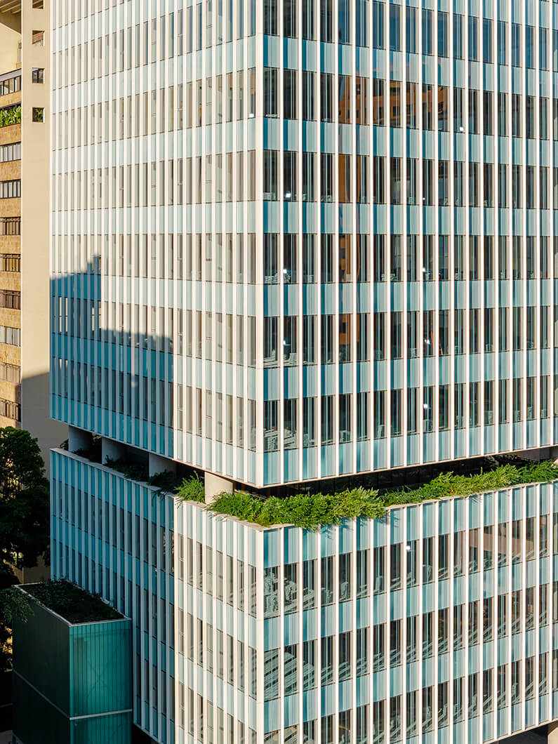 Aerial view of a glass-clad high-rise building featuring green terraces and vertical lines, surrounded by urban structures and greenery.