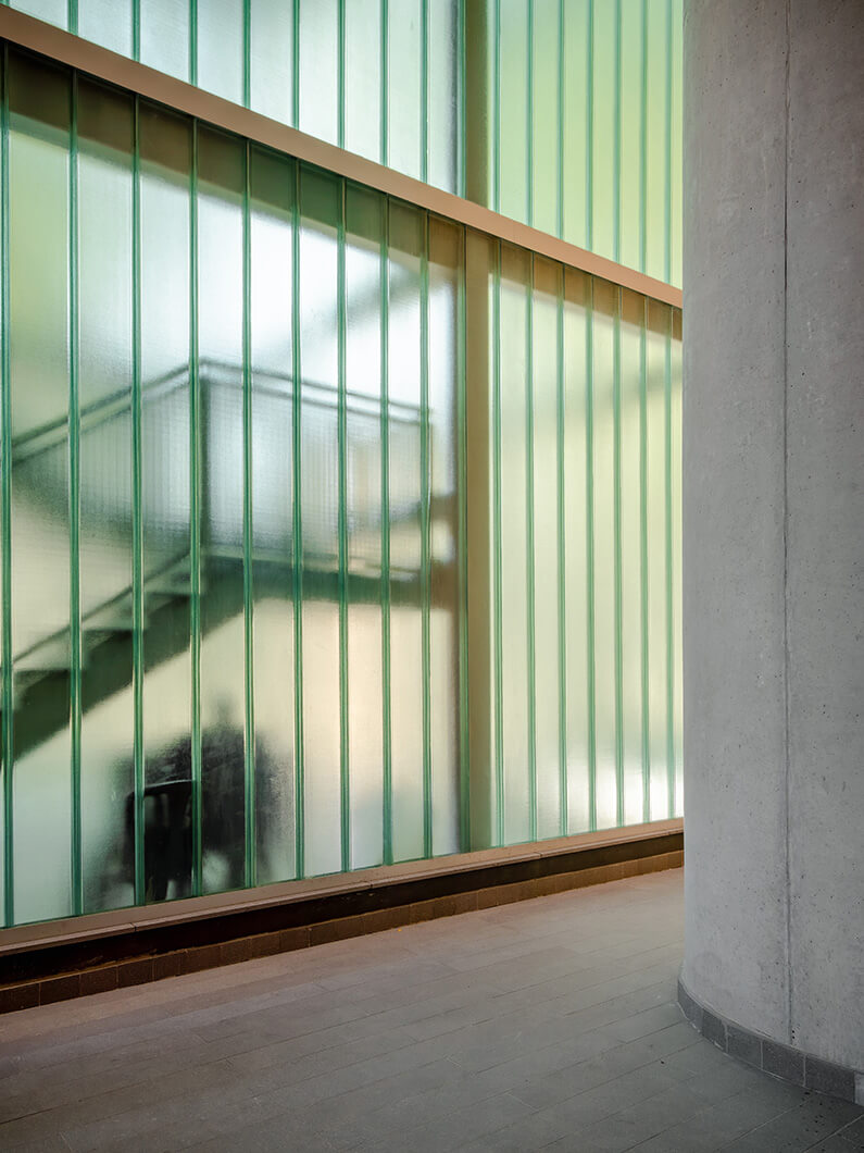 Interior shot featuring glass walls with vertical panels and a concrete column, showing an open staircase in the background.