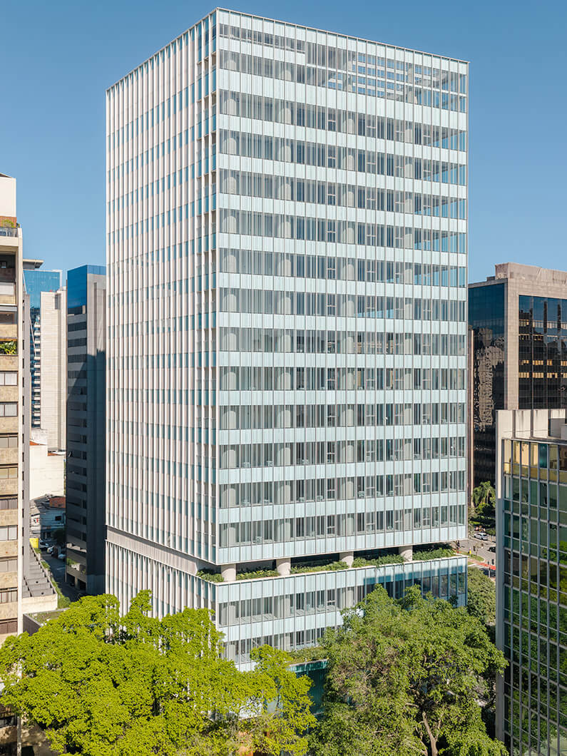 Glass-fronted office building surrounded by greenery in an urban setting, showcasing a sleek architectural design with multiple stories.