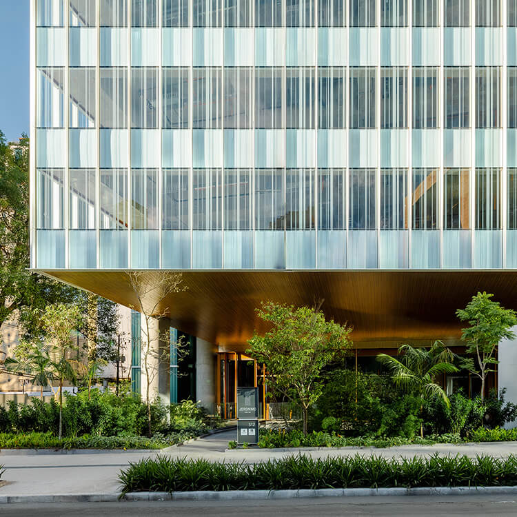 Glass-paneled building facade with greenery, showcasing architectural design. Entrance features a wooden overhang and landscaped surroundings.