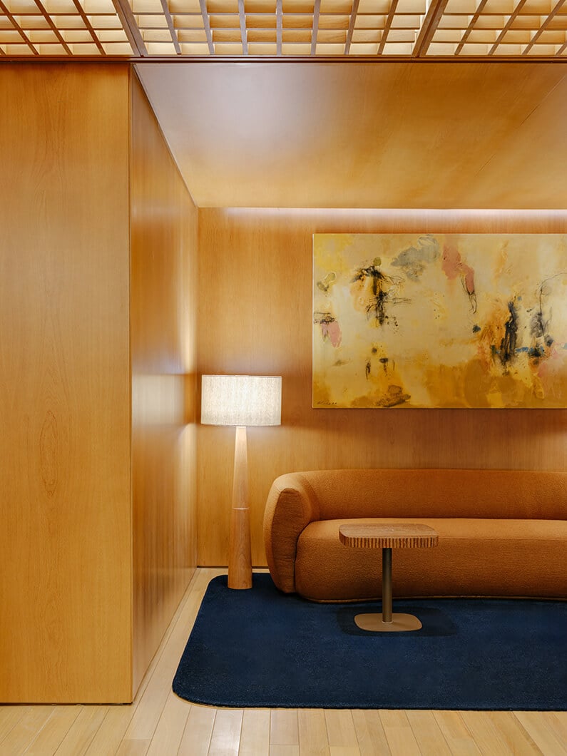 Cozy interior space featuring an orange sofa, wooden side table, floor lamp, and abstract wall art against warm wooden paneling.