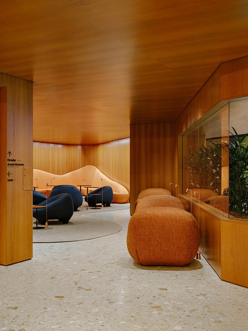 Interior view of a lounge area featuring curved seating in blue and orange, wooden walls, and a glass plant display. Soft lighting enhances the space.