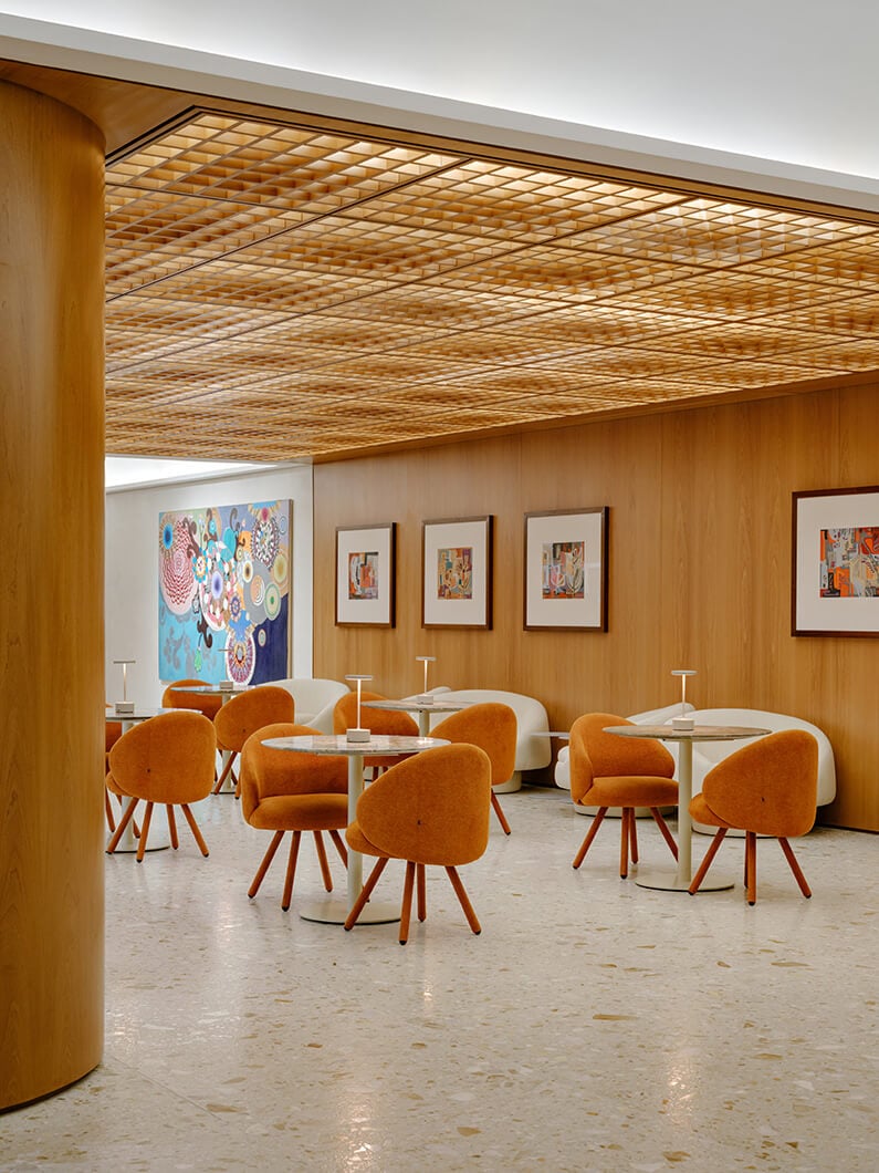 Interior of a cafe featuring wood paneling, orange chairs, circular tables, and colorful artwork on the walls. Soft lighting enhances the space.