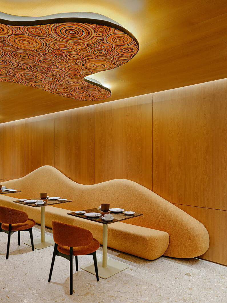 Interior of a restaurant featuring a unique ceiling design and curved seating in warm tones, along with simple tables and chairs set for dining.