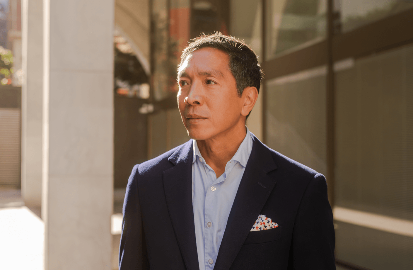 Businessman in a tailored suit stands in an urban setting, looking thoughtfully ahead under natural light.