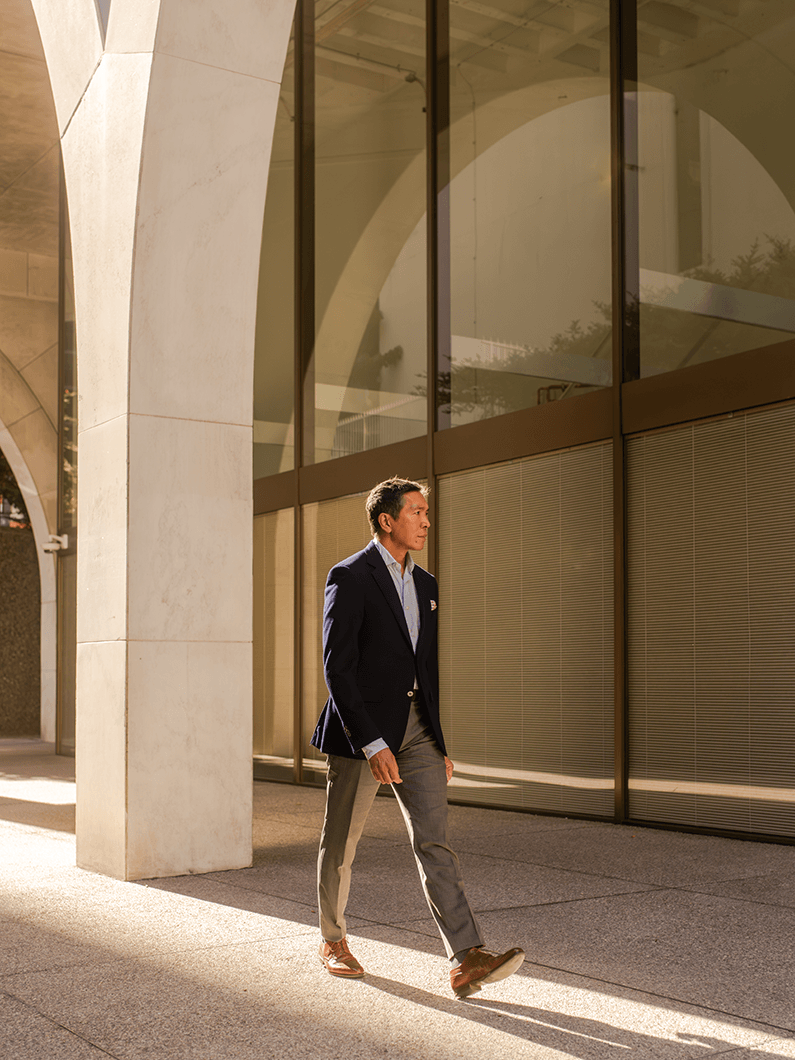 A man in a suit walks along a sidewalk under arched architecture, with large windows reflecting the sunlight.