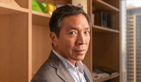 Business professional with short gray hair, wearing a suit, looking towards the camera in an office with wooden shelves and city view.